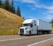 White semi truck with a trailer driving on a mountain highway under a blue sky, representing freight transportation and logistics services.