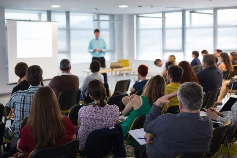 Business training seminar with presenter speaking to an audience in a modern conference room with projector and large windows