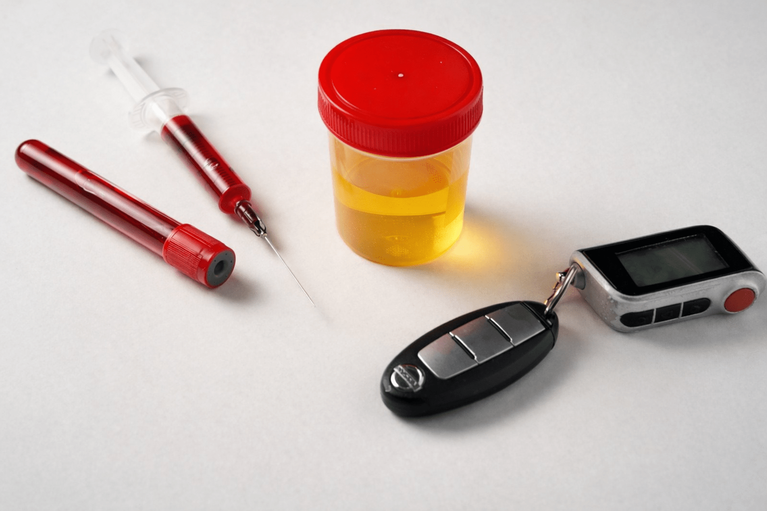 Drug and alcohol testing samples including blood vial, syringe, urine container, car keys, and breathalyzer device on a white background